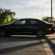 A person crouched beside a dark prestige sedan at the kerb photographing its paintwork with a phone, backlit in late-afternoon light