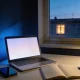 A desk at dusk with an open laptop showing a plain pale screen, a phone with a dark screen, a notebook of blank pages, a mug and a lamp