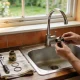 Hands working on a dismantled kitchen mixer tap over a stainless steel sink, the cartridge, O-rings, spanner and screwdriver laid out on a tea towel beside it