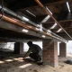 A person crouched under a raised timber-floored house with one hand laid along a copper pipe run, daylight falling between the floorboards above
