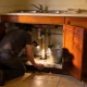 A person kneeling at an open sink cupboard with a torch, checking the waste trap and the cabinet floor for a damp patch