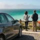 Two people standing at a coastal lookout railing with coffee cups beside a parked car, looking out over turquoise sea towards a headland