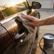 A hand wiping a dark car's door panel with a microfibre cloth, water beading on the paint, with a soapy bucket, hose and towels on the driveway