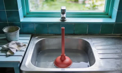 A stainless steel kitchen sink half full of still water with a red plunger standing in it, bucket and cloth on the bench beside it