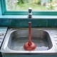 A stainless steel kitchen sink half full of still water with a red plunger standing in it, bucket and cloth on the bench beside it