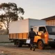 A white box truck with timber tray gates parked at a depot in dawn light, with the driver walking to the cab holding a clipboard
