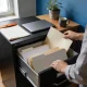 A person filing a plain folder into an open cabinet drawer in a home study, with a closed laptop, mug and plant on the desk beside them