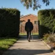 A person walking away along a suburban footpath with a walking stick, between clipped hedges towards a brick house in morning light