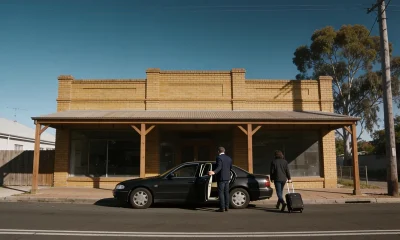 A plain sedan at the kerb of a country town shopfront with its rear door held open, a passenger with a suitcase walking towards it