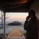 A person on the verandah of a country house at dawn with a phone to their ear, a plain car waiting at the kerb beyond a frosty garden