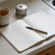 An open blank notepad with a pen on a kitchen bench, a mug of tea and a plain metal tin beside it, in soft winter morning light