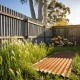 The back corner of a suburban Australian yard in spring sun, with long grass along a corrugated fence, an old sheet of roofing iron on the ground, a woodpile and gum trees beyond