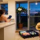 A woman at a kitchen bench holding a plain navy booklet, an open suitcase of summer clothes and a snorkel mask on the floor, a child in a straw sun hat on a chair and a potted frangipani by the glass door at dusk