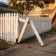 A white picket fence beside a concrete driveway with two pickets snapped at the base and leaning out, small shards of red plastic on the concrete, and a frangipani and the steps of a Queenslander behind in late sun