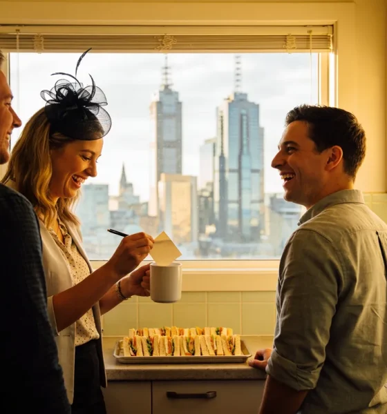 Three colleagues laughing in a small office kitchen, a woman in a black fascinator holding a paper slip over a white mug, a tray of sandwiches on the bench and city towers through the window