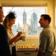 Three colleagues laughing in a small office kitchen, a woman in a black fascinator holding a paper slip over a white mug, a tray of sandwiches on the bench and city towers through the window