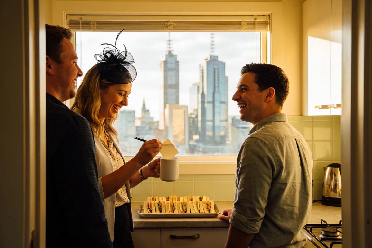 Three colleagues laughing in a small office kitchen, a woman in a black fascinator holding a paper slip over a white mug, a tray of sandwiches on the bench and city towers through the window