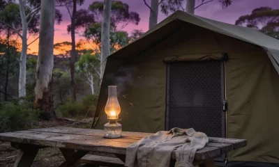 A canvas tent with its mesh door zipped shut, a glowing glass lantern on a weathered picnic table with a long-sleeved shirt draped over it, and a few mosquitoes in the light among tall gum trees at dusk