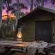 A canvas tent with its mesh door zipped shut, a glowing glass lantern on a weathered picnic table with a long-sleeved shirt draped over it, and a few mosquitoes in the light among tall gum trees at dusk