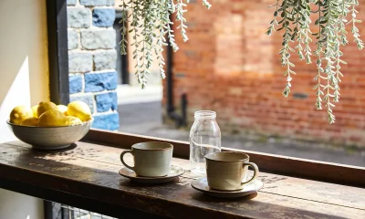 Two ceramic cups on saucers, an empty glass jar and a bowl of lemons on a worn timber counter in a Melbourne laneway cafe, with bluestone and red-brick walls behind in spring sun