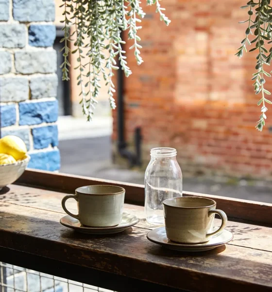Two ceramic cups on saucers, an empty glass jar and a bowl of lemons on a worn timber counter in a Melbourne laneway cafe, with bluestone and red-brick walls behind in spring sun