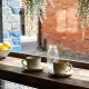 Two ceramic cups on saucers, an empty glass jar and a bowl of lemons on a worn timber counter in a Melbourne laneway cafe, with bluestone and red-brick walls behind in spring sun