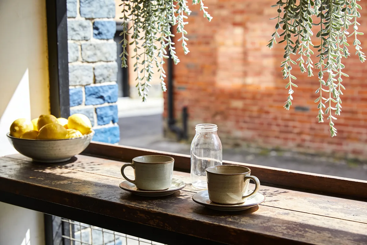 Two ceramic cups on saucers, an empty glass jar and a bowl of lemons on a worn timber counter in a Melbourne laneway cafe, with bluestone and red-brick walls behind in spring sun