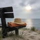 A straw hat on a folded orange beach towel on a dark timber bench above the sand on an overcast morning, with the sun breaking through grey cloud over a calm bay