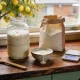 A glass jar of bubbly sourdough starter risen well above a rubber band, beside a bowl of poured-off starter, a paper bag of flour, kitchen scales and a wooden spoon on a timber bench by a window with a lemon tree outside